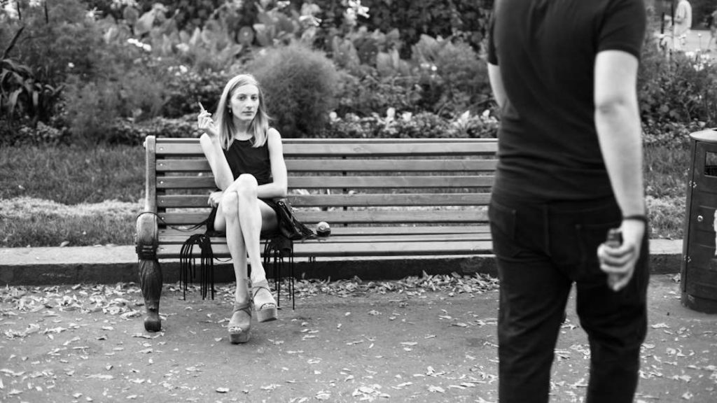 Une photo en noir et blanc d'une jeune femme en train de fumer une cigarette, assise sur un banc dans un parc.