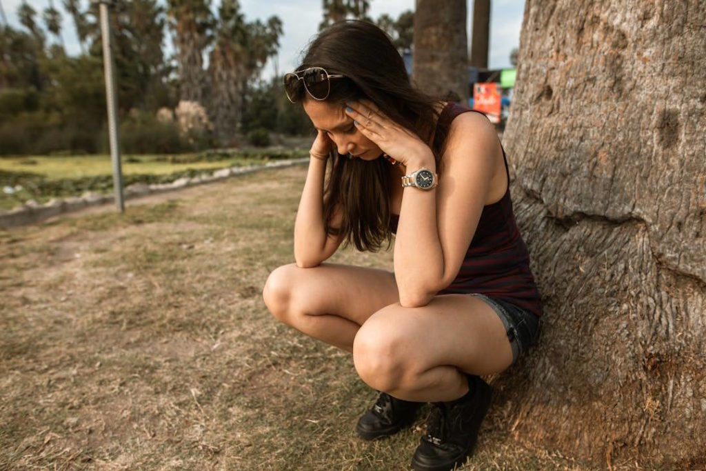 Une fille semble avoir mal au crâne, accroupie dehors contre un arbre.