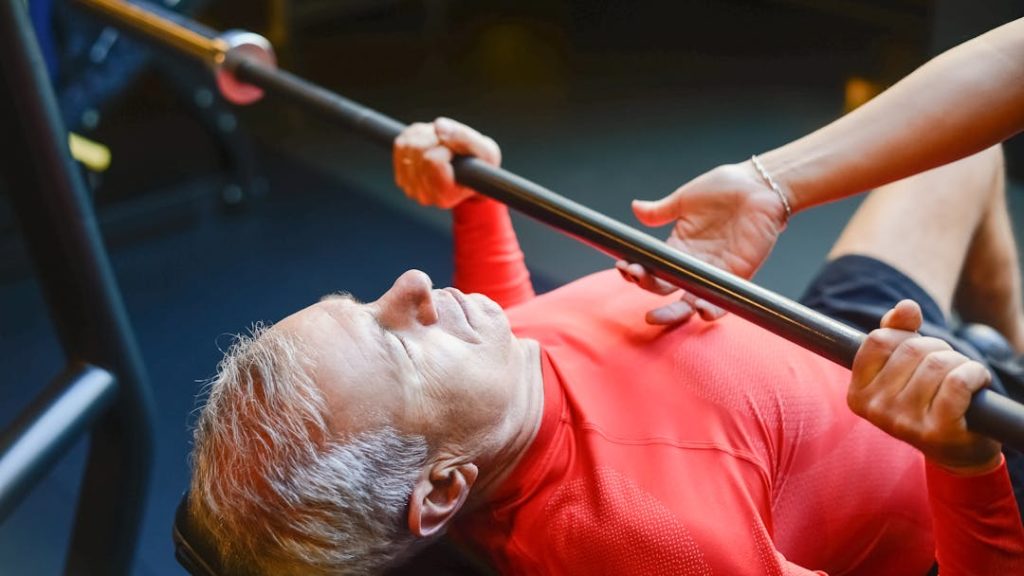 Un homme avec un t-shirt rouge, porte une barre avec un coach qui corrige sa posture.